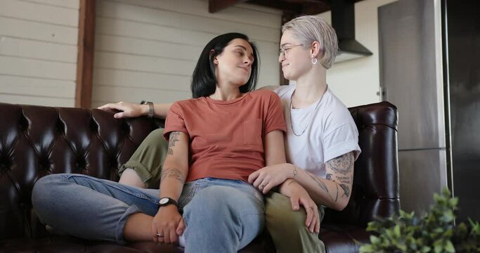 Joyful lesbian couple with black tattoos on arms talks smiling and hugs sitting on brown leather couch at home slow motion