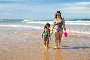 Happy mom and little girl wearing swimsuits, walking ankle deep in sea water on beach. Full length, front view. Family outdoor activities concept