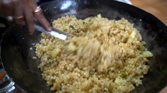 Person Stir And Mix Well The Sago Polenta With Potato Using A Stainless Flat Ladle In A Black Metal Kadai. - Close Up Shot
