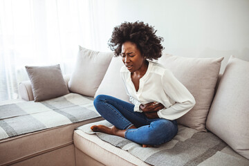Fototapeta premium Sad beautiful woman sitting on the couch and feeling spasm and symptoms of pms. Shot of a young woman experiencing stomach pain on the sofa at home