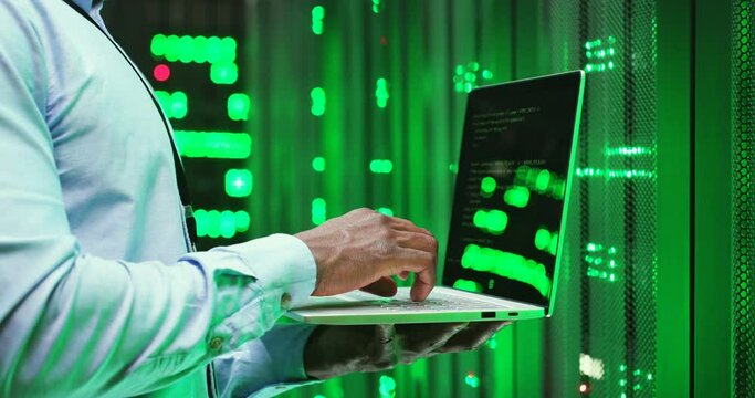 Close up of African American man standing among servers with laptop computer, typing on keyboard and checking big data processors. Male analytic in data storage working on digital information security