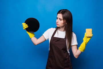 Young woman in apron with sponge looking at washed dish on blue background. Clean dishes, order in...