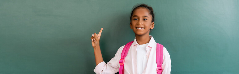 Horizontal concept of african american schoolgirl pointing at green chalkboard in classroom - Powered by Adobe