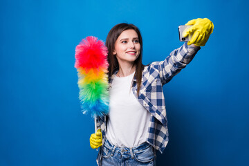 Joyful woman in yellow rubber gloves for hands protection taking selfie on mobile phone while holding colorful duster brush during cleaning isolated over blue background