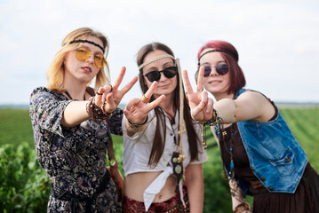 Three hippie women, wearing boho style clothes, showing peace sign on green currant field, having fun. Female friends, traveling together in countryside. Eco tourism concept. Summer leisure free time
