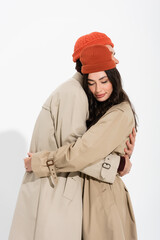 stylish couple in beanie hats and trench coats hugging on white