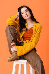 trendy woman in autumn outfit sitting on white stool and looking at camera isolated on orange