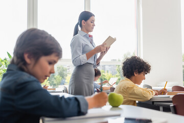 selective focus of teacher reading book while multicultural pupils writing during lesson