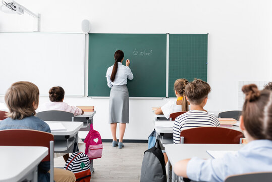back view of teacher in formal wear writing on chalkboard near multiethnic pupils in classroom
