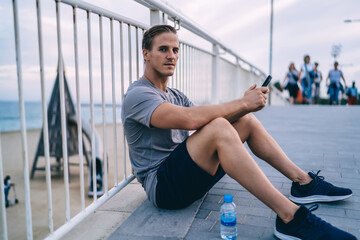 Obraz premium Portrait of handsome male athlete resting after workout using smartphone for chatting and sending mails,sportsman looking at camera holding mobile phone browsing via 4G connection on break