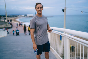 Half length portrait of cheerful male athlete in active wear sending mail and chatting via mobile phone, happy sportsman looking at camera resting after training holding smartphone connected to 4G