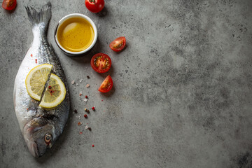 Fototapeta na wymiar Raw fish dorado with lemon, cut cherry tomatoes, olive oil, seasonings on gray concrete background ready for cooking. Whole uncooked fish and ingredients, healthy dinner or diet meal, space for text