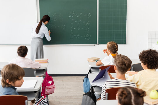 back view of teacher writing equations on chalkboard near multicultural pupils during math lesson - Powered by Adobe