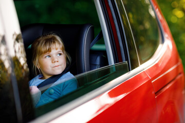Adorable toddler girl sitting in car seat, holding plush soft toy and looking out of the window on...