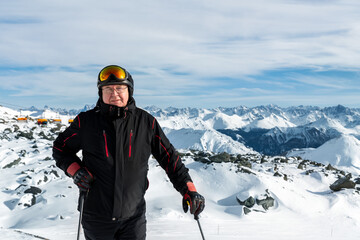 Active senior experienced mature old person in ski helmet, goggles and black suit stand on mountain peak enjoy winter extreme sport activities. Man on stunning panoramic mountains sunshine background