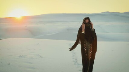 A beautiful young woman in a black oriental dress walking in the desert. The head is covered with a scarf, the face is hidden. Arabic nature background, white sands, dramatic sky and sun at sunset - Powered by Adobe