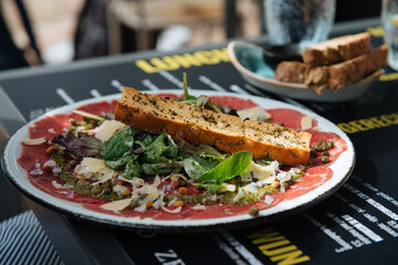 A plate with Beef Carpaccio, with cheese, lettuce, pine nuts, delicious with a sandwich, stands on a terrace table, people eat and drink in the background