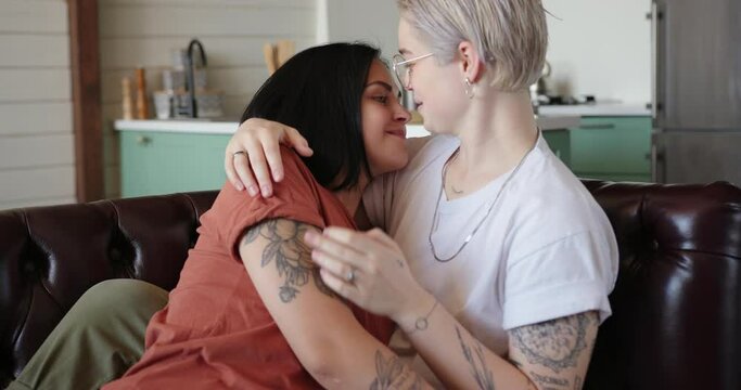Dark haired lesbian in orange t-shirt massages girlfriend neck and kisses sitting on leather sofa at home slow motion closeup
