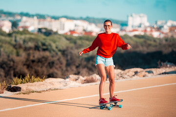 Sporty cheerful smiling woman in blue shorts, red sneakers and hoodie riding skateboard in sunny summer day. Vacation activities