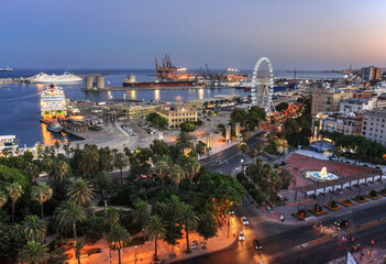 Malaga, Spain: seafront and harbour at sunset  with ferris wheel at the harbour entrance. In the background the harbour in full operation with a freighter and harbour cranes.