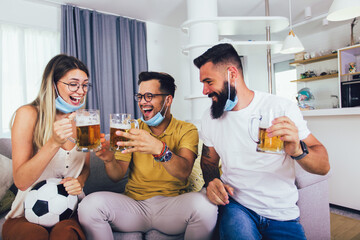 Group of young friends watching a football match at home, cheering and drinking beer, wearing protective mask.