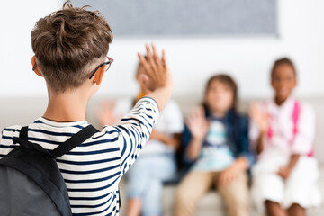 Obraz premium Selective focus of schoolboy with backpack waving hand at multiethnic classmates