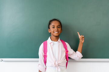 African american schoolchild with backpack looking at camera while pointing with finger near chalkboard