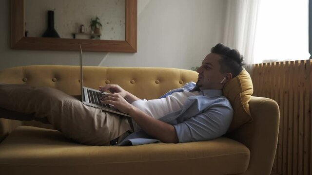 A young man is lying on the sofa with a computer on his lap and working