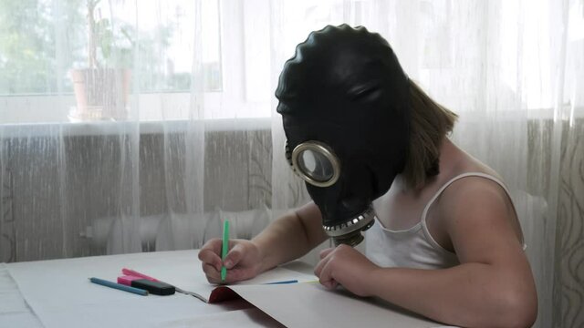 strange, weird little girl in gas mask sitting at table, drawing illustration with multicolored felt pens in album. protection during covid-19 pandemic quarantine, stay home funny concept