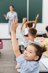 selective focus of schoolgirl with hand in air looking at camera near classmates and teacher on...