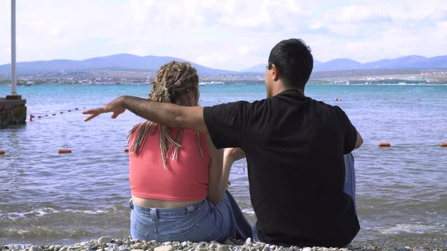 Couple hugging and drinking wine on beach. Media. Young cute couple relaxing sitting on rocky beach. Couple in love on vacation look at seascape with mountains
