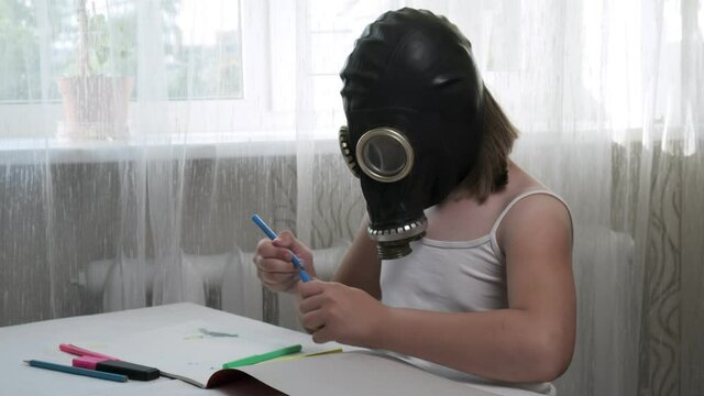 strange, weird little girl in gas mask sitting at table, drawing illustration with multicolored felt pens in album. protection during covid-19 pandemic quarantine, stay home funny concept