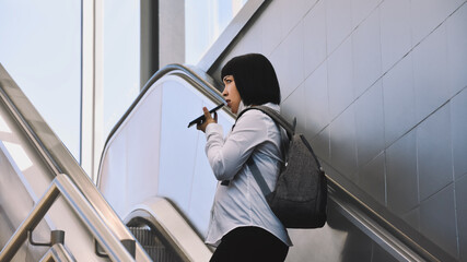 A young oriental woman talks on her smartphone with her face mask down while using an escalator of public transport.