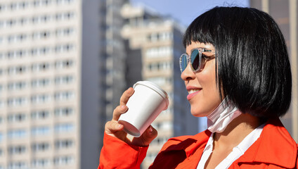 A young asian woman with sunglasses and her face mask removed brings a hot drink to her lips, enjoying the morning sun on a blurred midtown background.