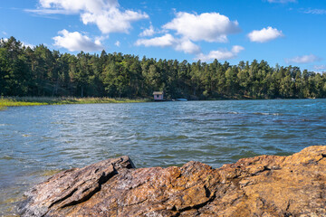 Stony shore of the Baltic Sea on a sunny autumn day. Big granite rock on the foreground. Green forest islands of Scandinavia. Beautiful Swedish landscape seaside with blue sky and white clouds.