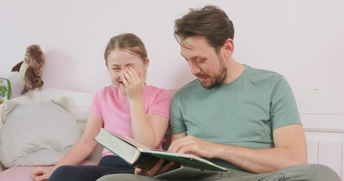 Father and teen daughter are sitting on the sofa, and are looking at the photo album. The girl laughs and wipes the eye.