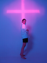 Cool young girl standing in blue neon light with pink cross in studio. Trendy poster to announce party 