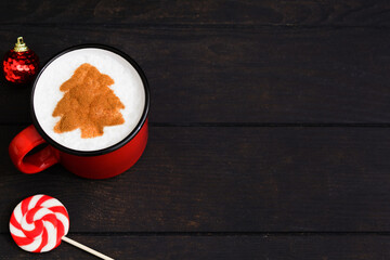 Christmas red cup of coffee with fir tree pattern. New Year beverage over dark wooden background with copy space. Mag of cuppuccino with art in shape of Christmas tree and lollipop
