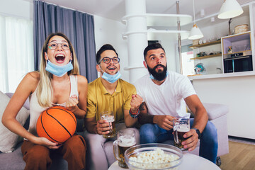 Happy friends or basketball fans watching basketball game on tv at home and celebrating victory at home, wearing protective mask.Friendship, sports and entertainment concept.