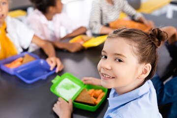 Obraz premium selective focus of schoolgirl looking at camera near lunch box with sandwich and fresh carrots