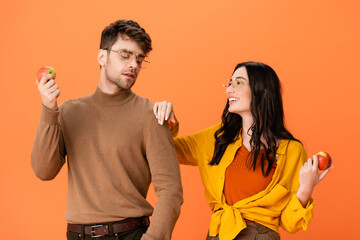 stylish couple in glasses and autumn outfit holding apples while looking at each other isolated on orange