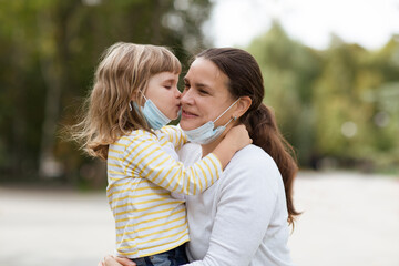 Mom and child girl with medical face lowered mask hugging outdoors
