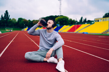 Young caucasian male in active wear and headphones having break on training drinking water from bottle, thirsty sportsman in sound accessory making pause for refreshment on workout on stadium