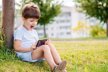 Cheerful child sitting on the grass looks cartoons in the phone in the summer at sunset. Cute boy having fun in nature