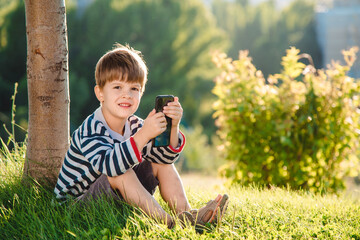 Fototapeta premium Cheerful boy sitting on the grass looks cartoons in the phone in the summer at sunset. Cute baby having fun in nature