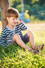 Naklejka na ściany i meble Cute boy sitting on the grass speaks by phone in the summer at sunset. The child communicates on a mobile