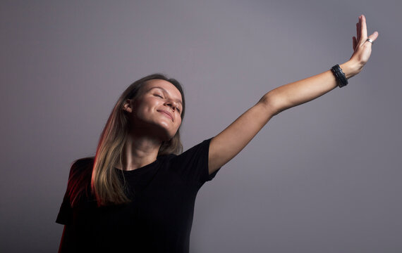 headshot of a young glad calm caucasian woman with closed eyes with satisfied expression waking up streaching her hand to the light. dark grey studio background, casula clothes