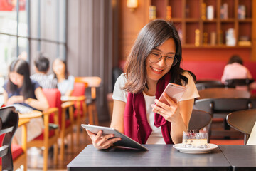 Lifestyle, Technology Concept - Asian woman typing write message on smart phone and using tablet sitting at a table with cake in a modern cafe.