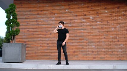 Wide frame with a handsome young man who uses his mobile phone to go with friends at a distance, during the pandemic. Caucasian young man in mask talking on the phone. Brick background. - Powered by Adobe