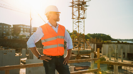 Confident Bearded Head Civil Engineer-Architect in Sunglasses Stands on a Construction Site on a Sunny Bright Day. Man is Wearing a Hard Hat, Shirt, Jeans and an Orange Safety Vest. 